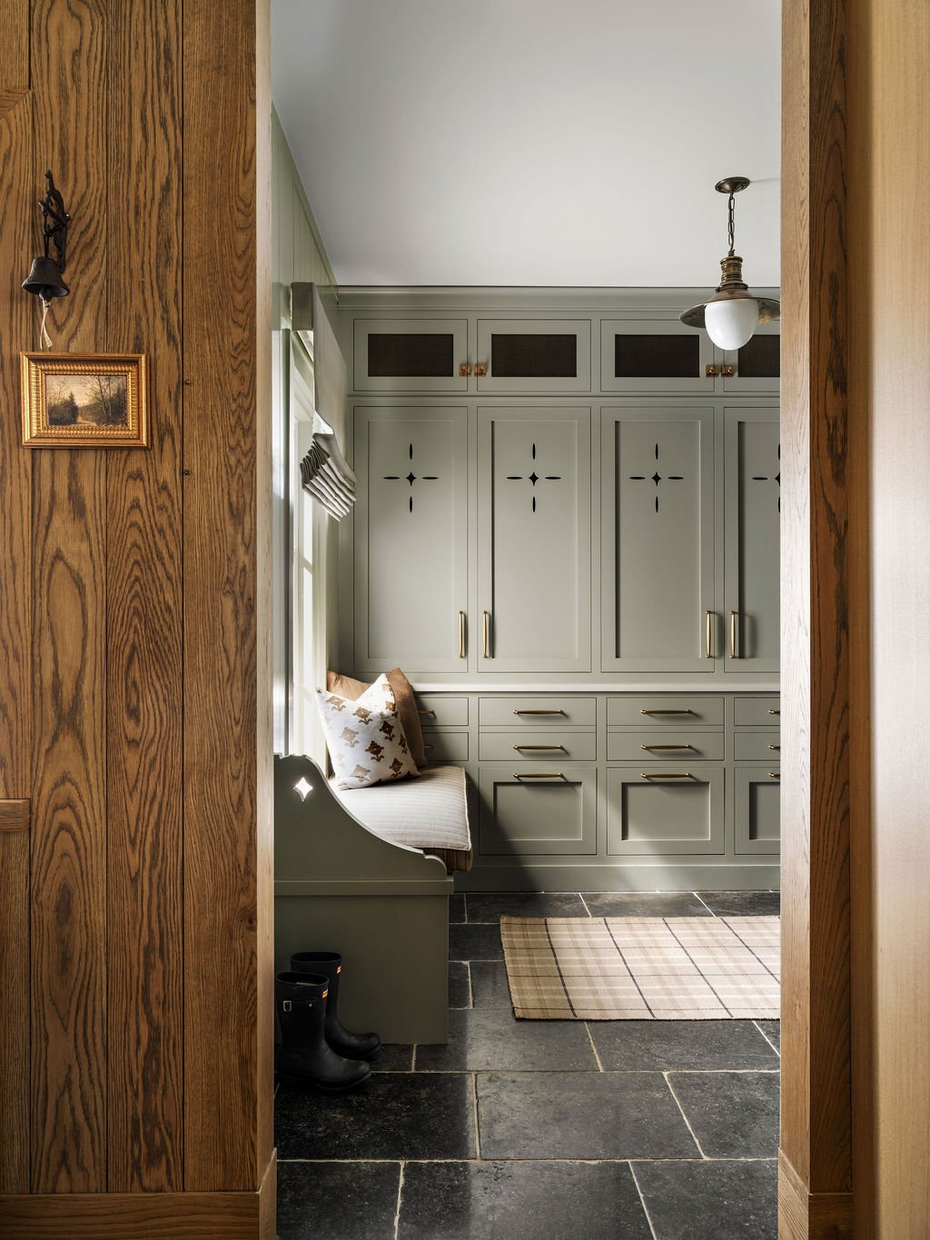 View into a cozy mudroom with green cabinetry, a built-in bench with pillows, and black rubber boots on the floor.