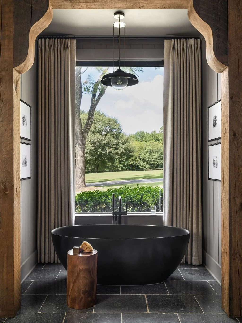 A bathroom with a black freestanding bathtub positioned in front of a large window with beige curtains. Outside view of trees and greenery. A hanging ceiling light, wooden side table with bath items, and framed pictures on the walls.