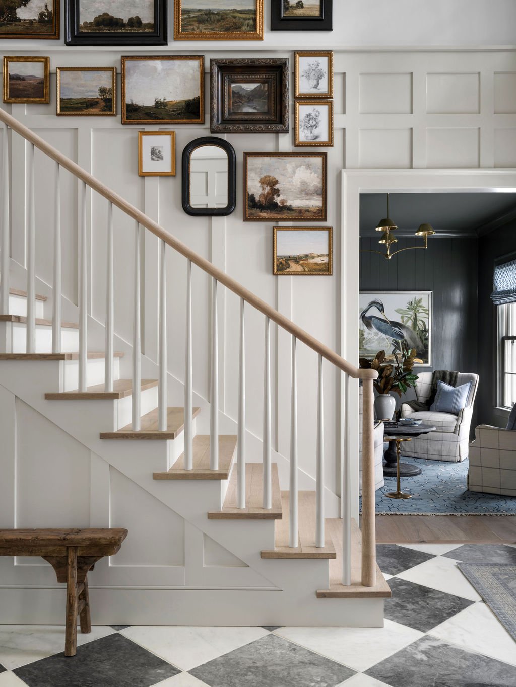 Interior view of a staircase hall with white painted panel walls decorated with a gallery wall of landscape paintings in various frames. To the right, a living room with a black accent wall, a large painting of a heron, and furniture including armcha