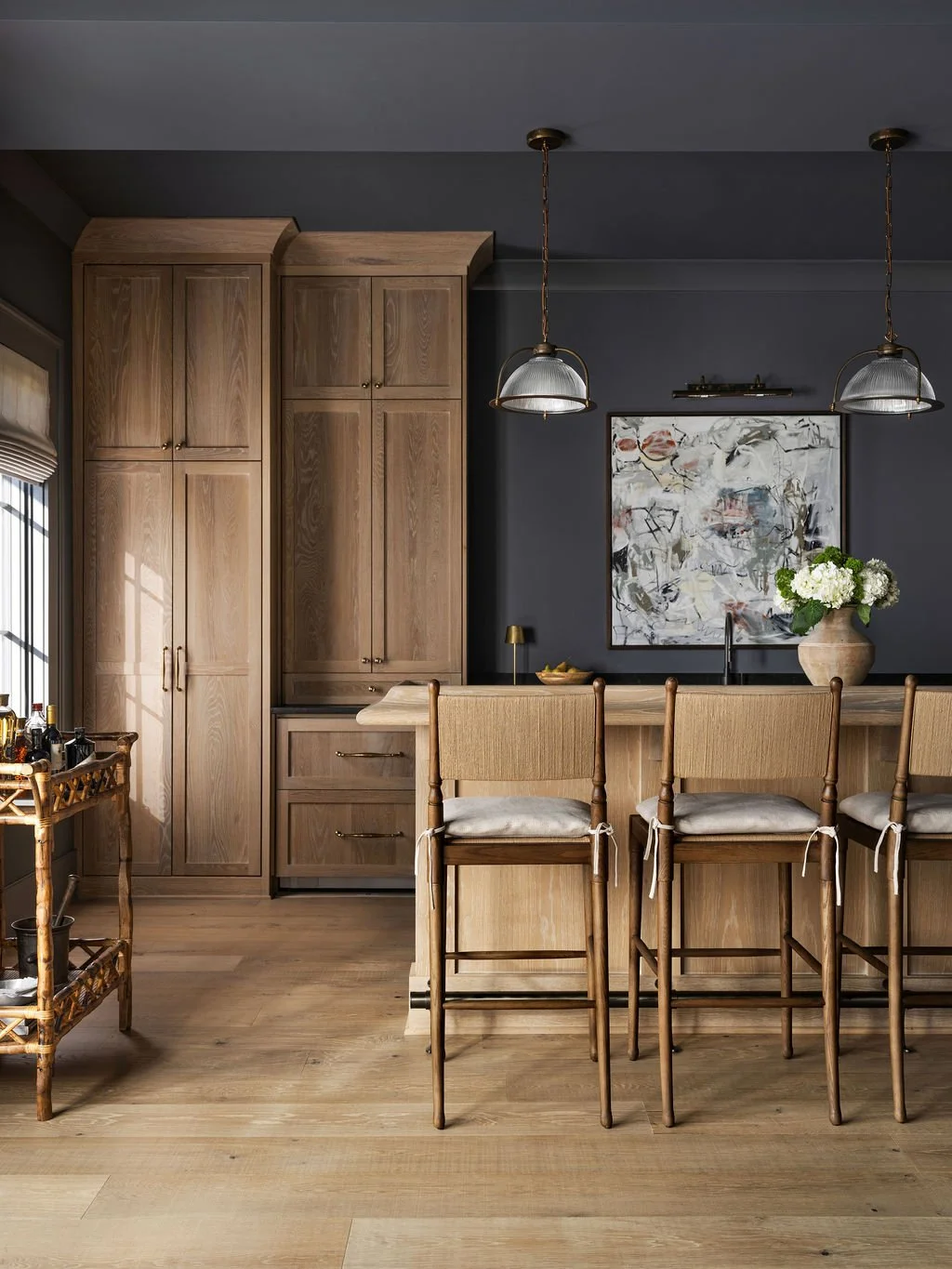 Kitchen with dark painted walls, wooden cabinets, a bar area with four wooden chairs, a large abstract painting, pendant lights, and a vase with white flowers.