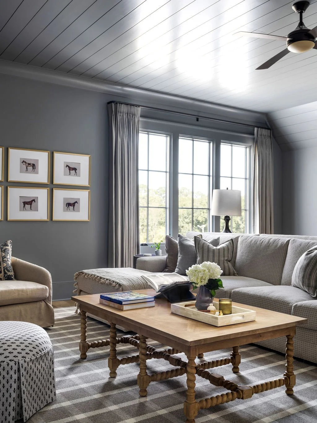 A cozy living room with a light-colored sofa and matching armchair, a wooden coffee table with books and a vase of white flowers, large windows with curtains, striped carpet, a wall with framed pictures of horses, a table lamp, and a ceiling fan.