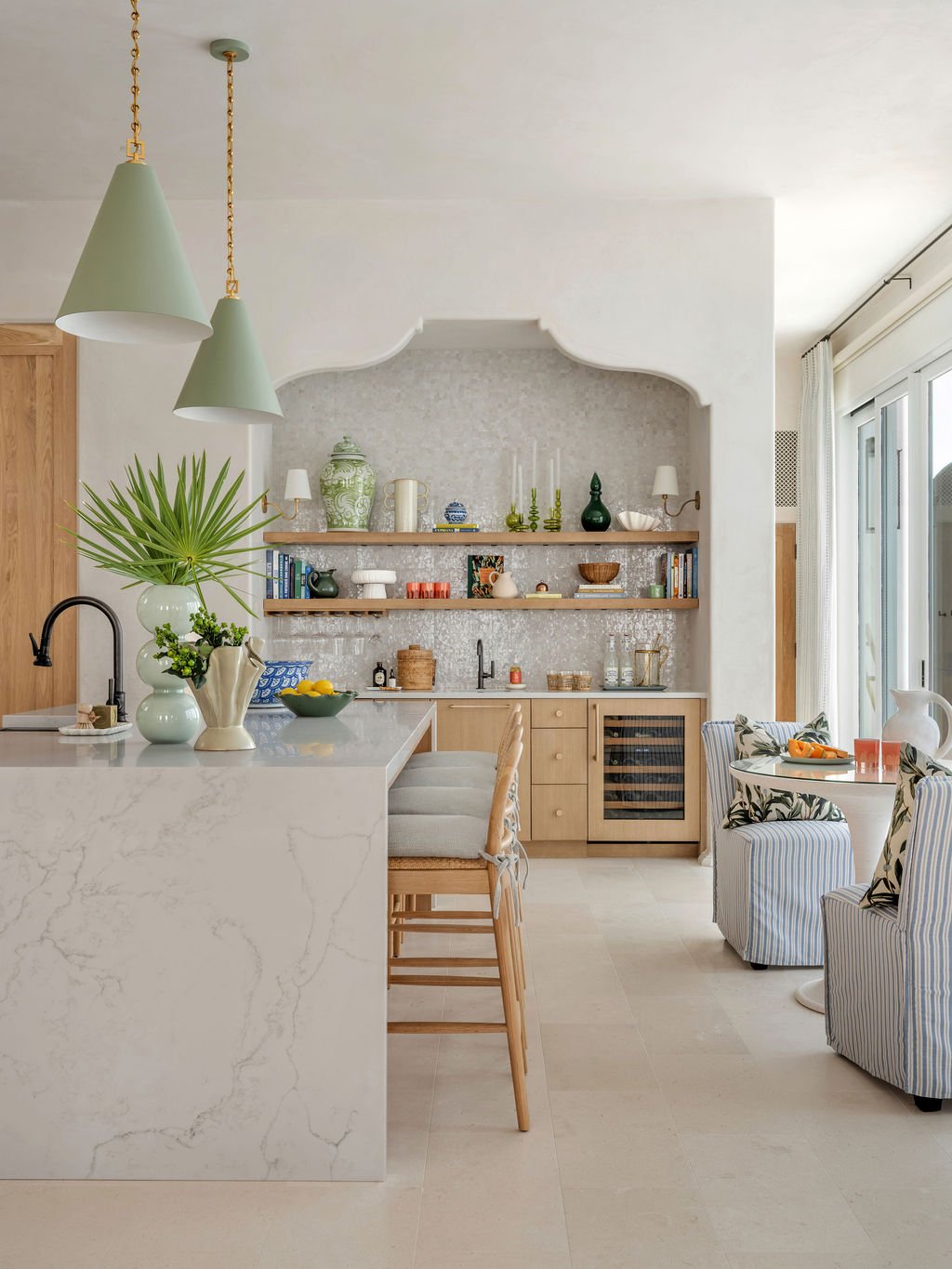 A modern kitchen with a white marble island, light wood cabinetry, and open shelves displaying decorative items. There are two dining chairs and a striped armchair with a small round table set for a meal. Large windows with curtains allow natural light into the space.