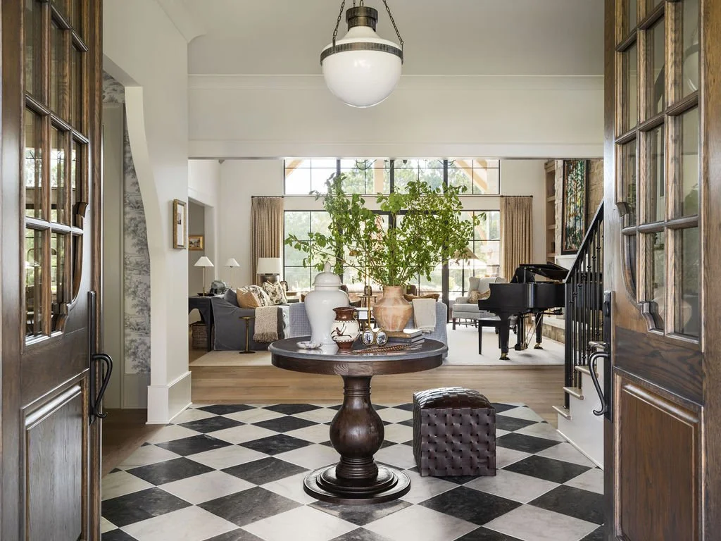 View of a spacious living room with a grand piano, large windows, and a round table with a large vase of green plants, seen through a doorway with wooden double doors.