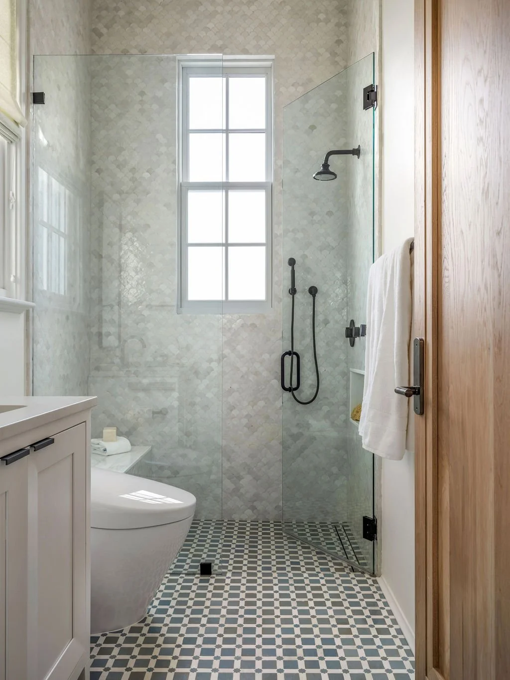 Modern bathroom with a glass-enclosed shower, black fixtures, a window, and a white toilet next to a wooden door.