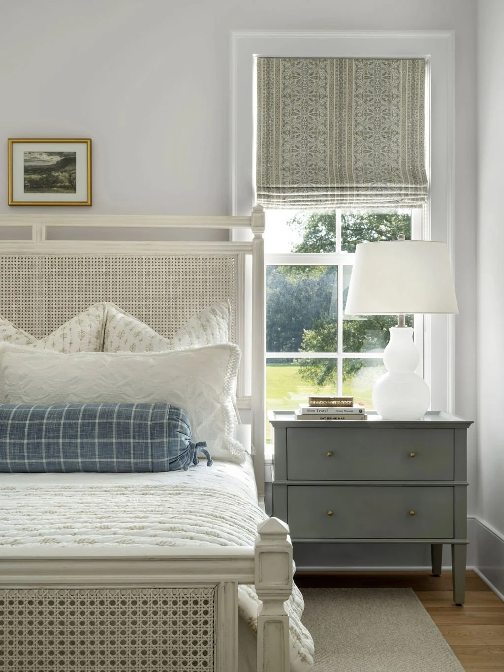 A cozy bedroom with white walls, a large window with patterned Roman shades, a white bed with a rattan headboard, and a gray nightstand holding a white lamp and books.