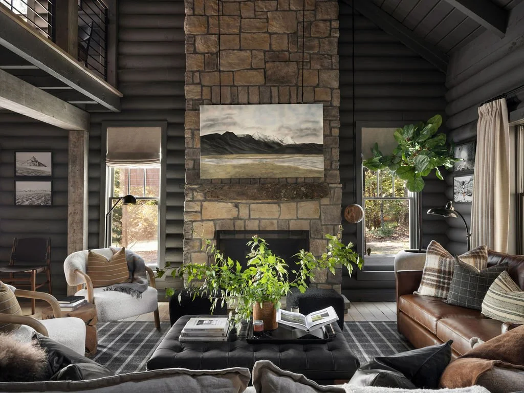Living room with a stone fireplace, black walls, large windows, and cozy furniture including a leather sofa, armchairs, and a black ottoman with books and a plant on it.