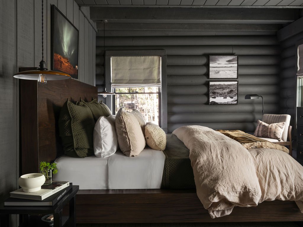 Bedroom with made bed, pillows, nightstand, and window with blinds, log cabin interior, wall-mounted TV, wall art.