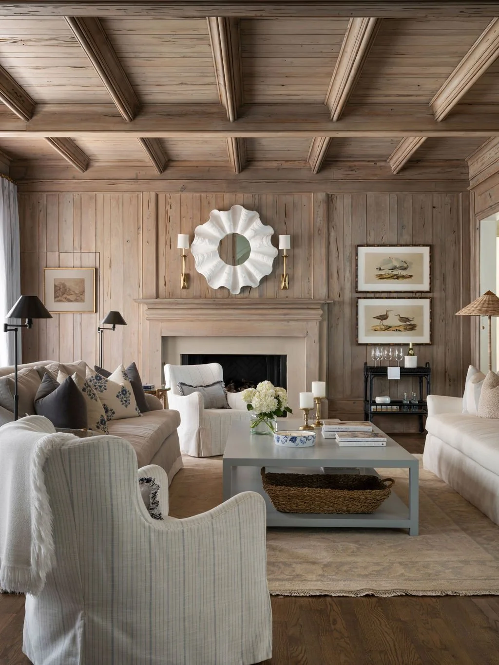 Cozy living room with wood-paneled walls and ceiling, featuring a fireplace, white and beige furniture, decorative pillows, framed artwork, a white coffee table with candles and a flowers, and various lamps and wall sconces.
