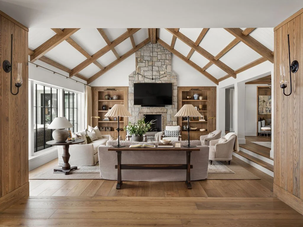 Living room with vaulted ceiling, stone fireplace, mounted TV, wooden shelves, beige sofas, armchairs, lamps, and large window
