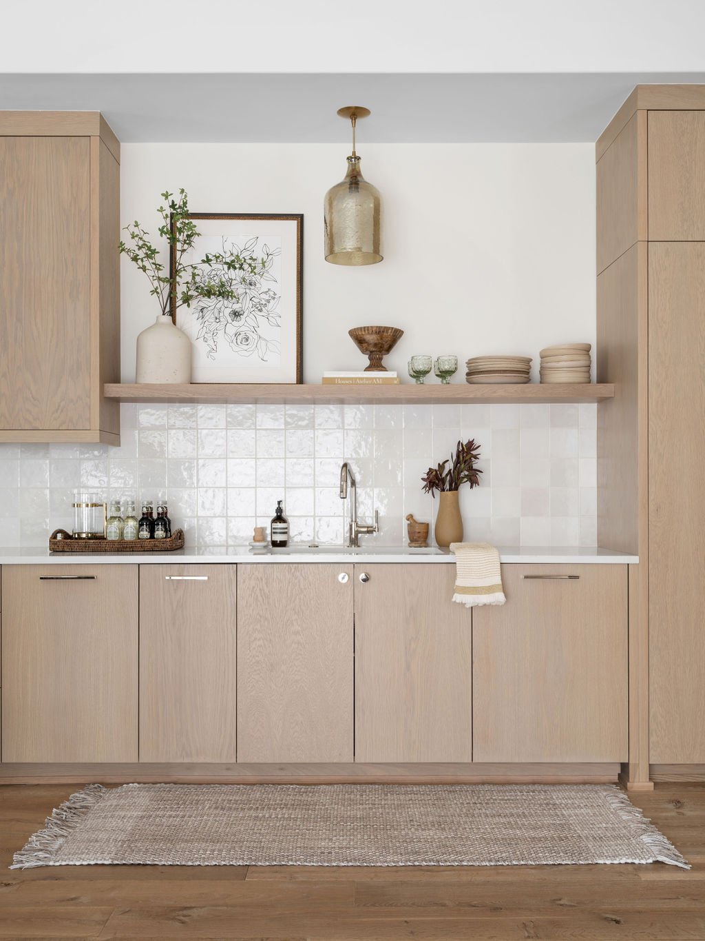 Minimalist kitchen with light wood cabinetry, white tiled backsplash, and open shelving with decorative items like a vase with greenery, plates, glasses, and artwork, along with a woven rug on wooden floor.