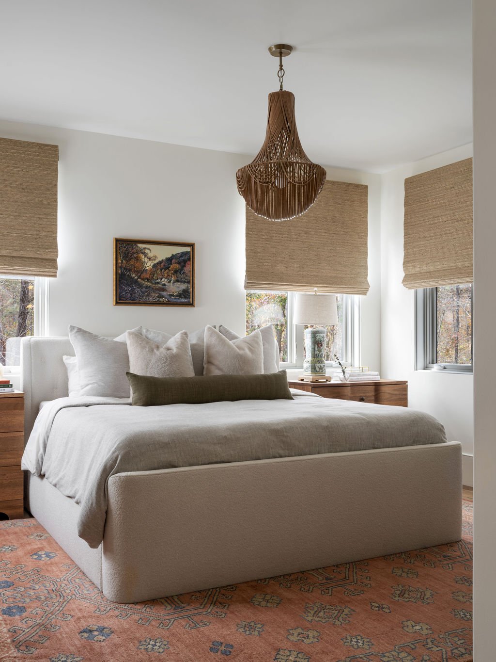 A bedroom with a bed, multiple pillows, nightstands, a lamp, a painting, and beige window blinds, with a peach-colored patterned rug on the floor.