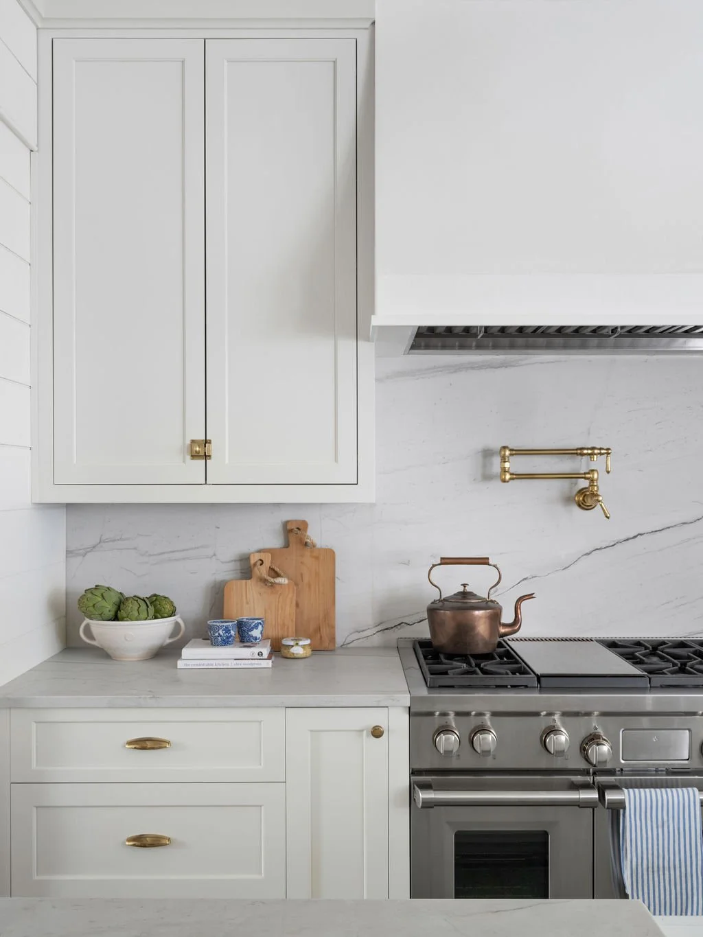 A modern kitchen with white cabinets, a marble countertop, brass fixtures, a kettle on a stainless steel stove, and artichokes in a bowl.
