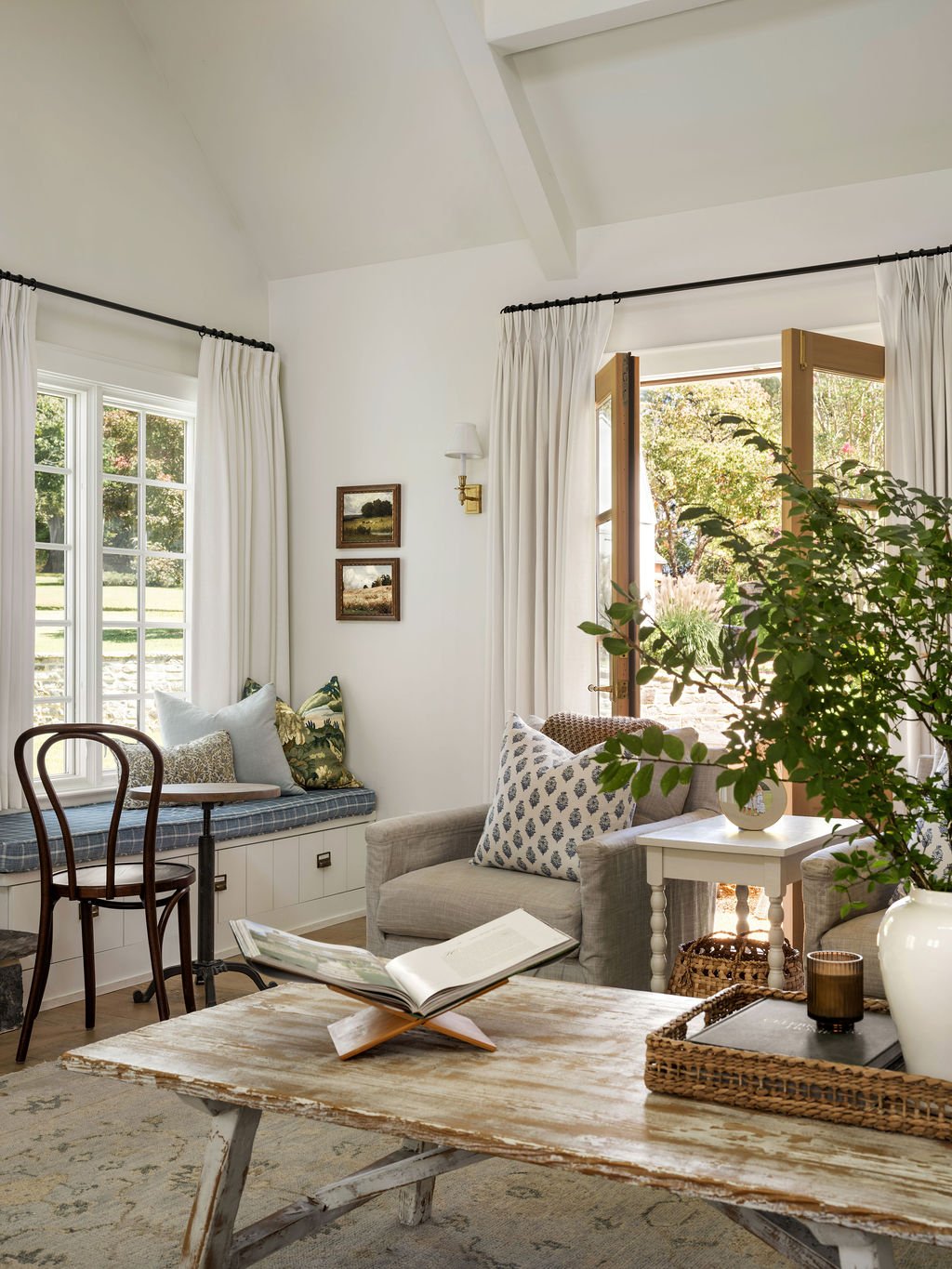 Cozy living room with large windows and open French doors, white curtains, potted plant, beige couch with decorative pillows, wooden coffee table with open book, additional seating nook with a window seat, art on the wall, and natural light.