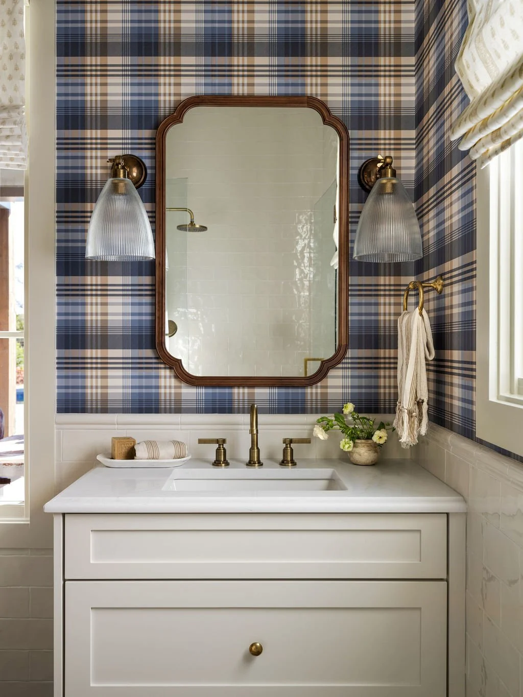 Bathroom vanity with white cabinet, marble countertop, gold fixtures, a mirror, wall-mounted light fixtures, a towel ring with a white cloth, and a small plant with white flowers. The wall has blue plaid wallpaper, and a window with a white frame and