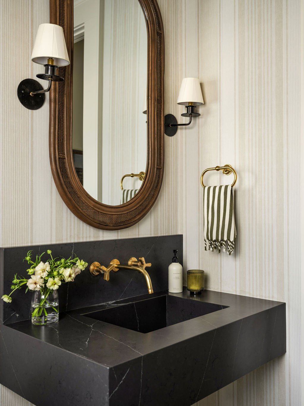 Bathroom with black marble sink, gold faucet, striped towel on gold towel ring, wall-mounted sconces with white shades, ornamate oval mirror, vase with white flowers, soap dispenser, and candle on a beige patterned wall.