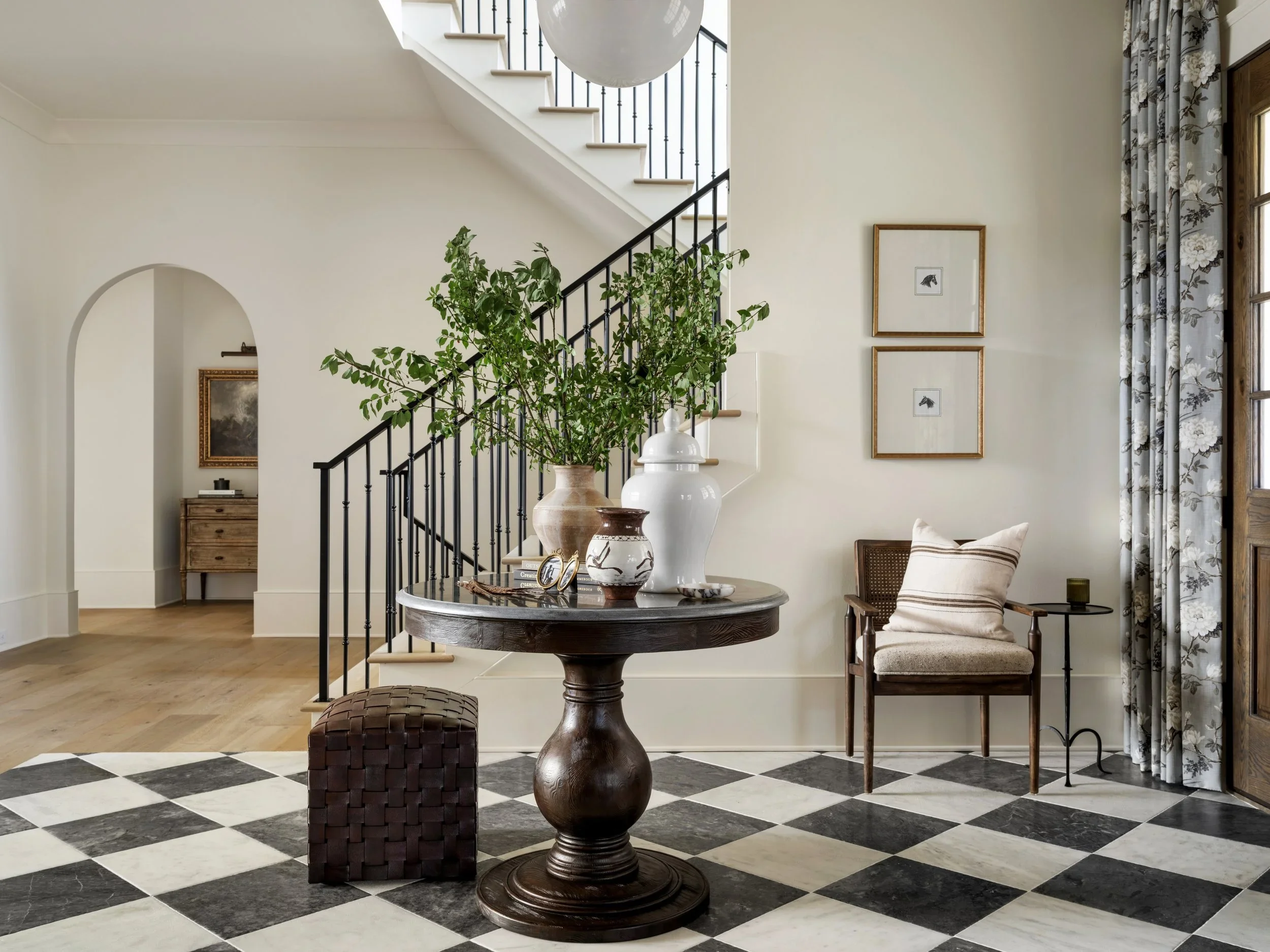 A living room with a checkered black and white marble floor, a round wooden table with vases and framed photos, a chair with a beige pillow, a large plant, framed wall art, and a staircase with black wrought iron railing leading upstairs, with a larg