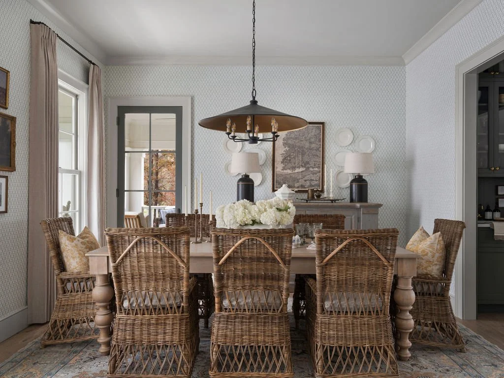 A dining room with a large wooden table surrounded by wicker chairs, two lamps on a sideboard, and a floral centerpiece. There are windows and a door leading outside, with pastel curtains and decorative plates on the wall.