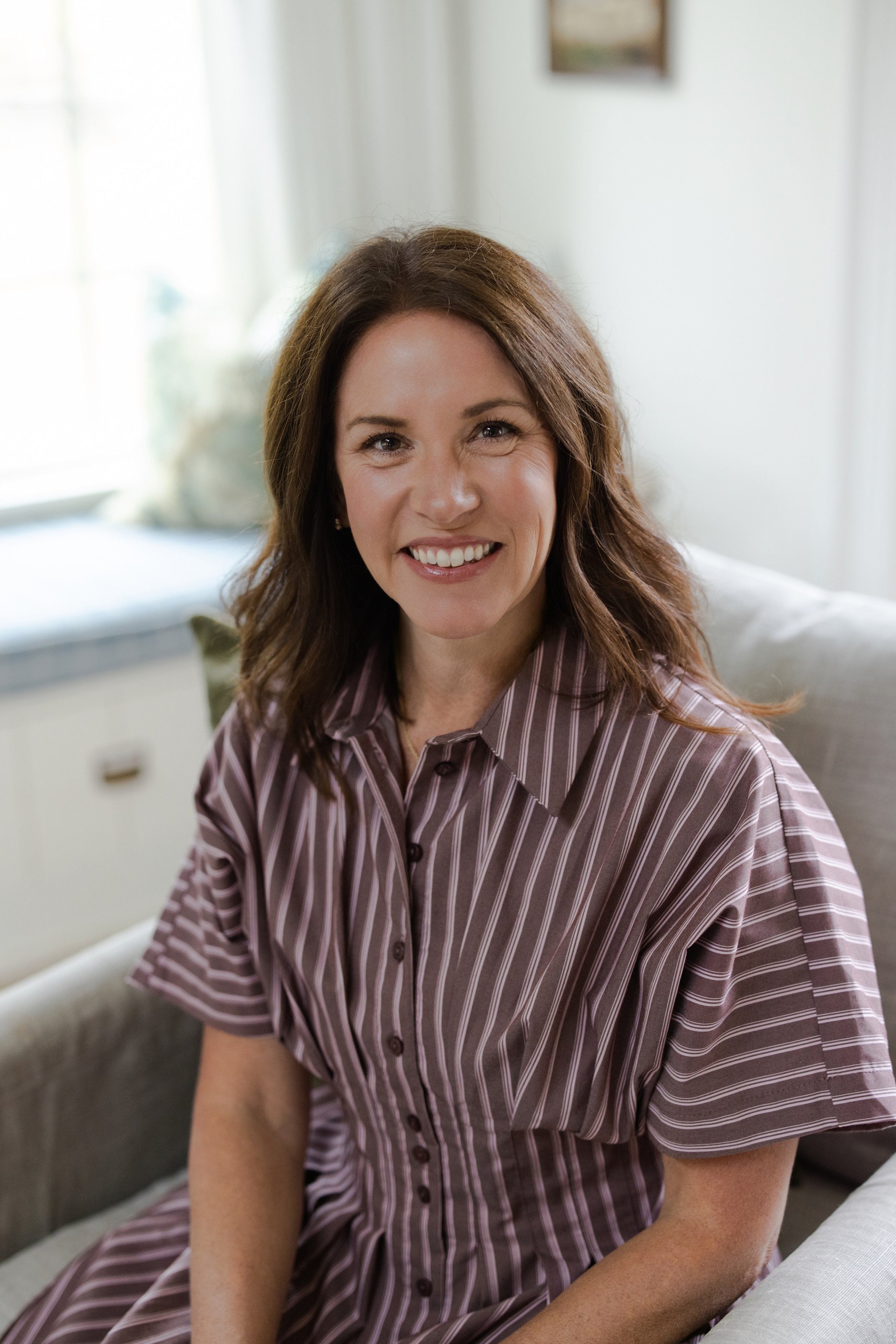 A woman with shoulder-length brown hair, wearing a striped button-up dress, sitting on a sofa in a bright room, smiling at the camera.