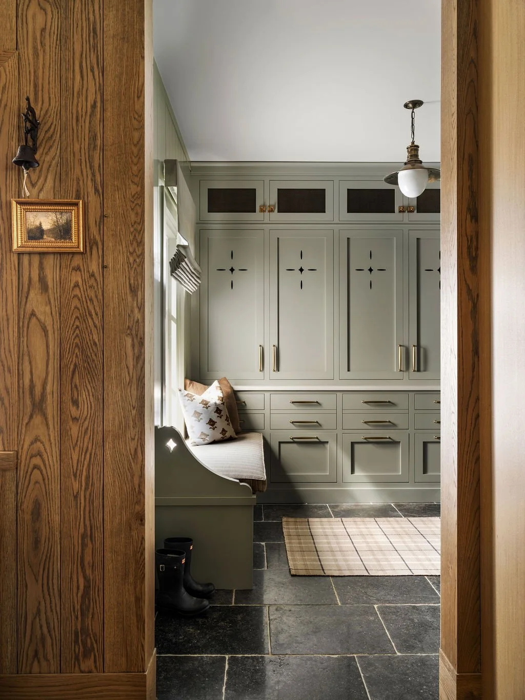 A cozy mudroom with built-in gray cabinets, a window seat with cushions, black rubber boots, a plaid rug on black stone tile floor, and a vintage-style ceiling light fixture.