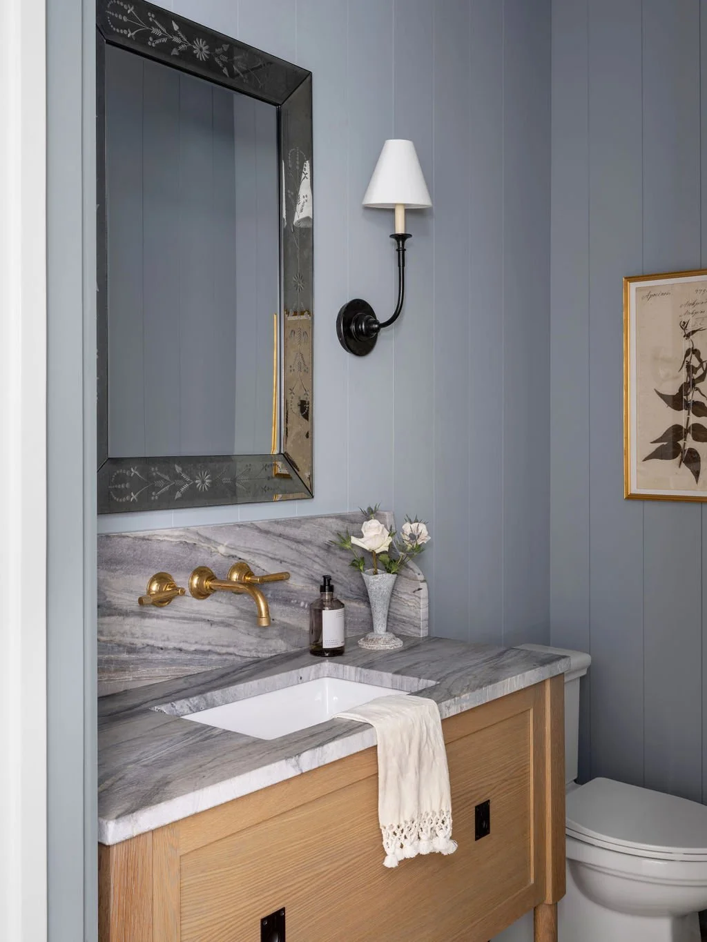 Bathroom with a marble countertop, a mirror with a decorative frame, a black and white wall sconce, and a vase with white flowers. A towel hangs over the sink and there's a framed botanical print on the wall.