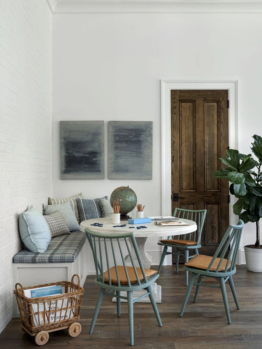 A cozy dining nook with a white round table, light blue chairs, a built-in bench with striped and checked pillows, and a large potted plant. There is a wooden door, framed abstract art, and a globe on the table.