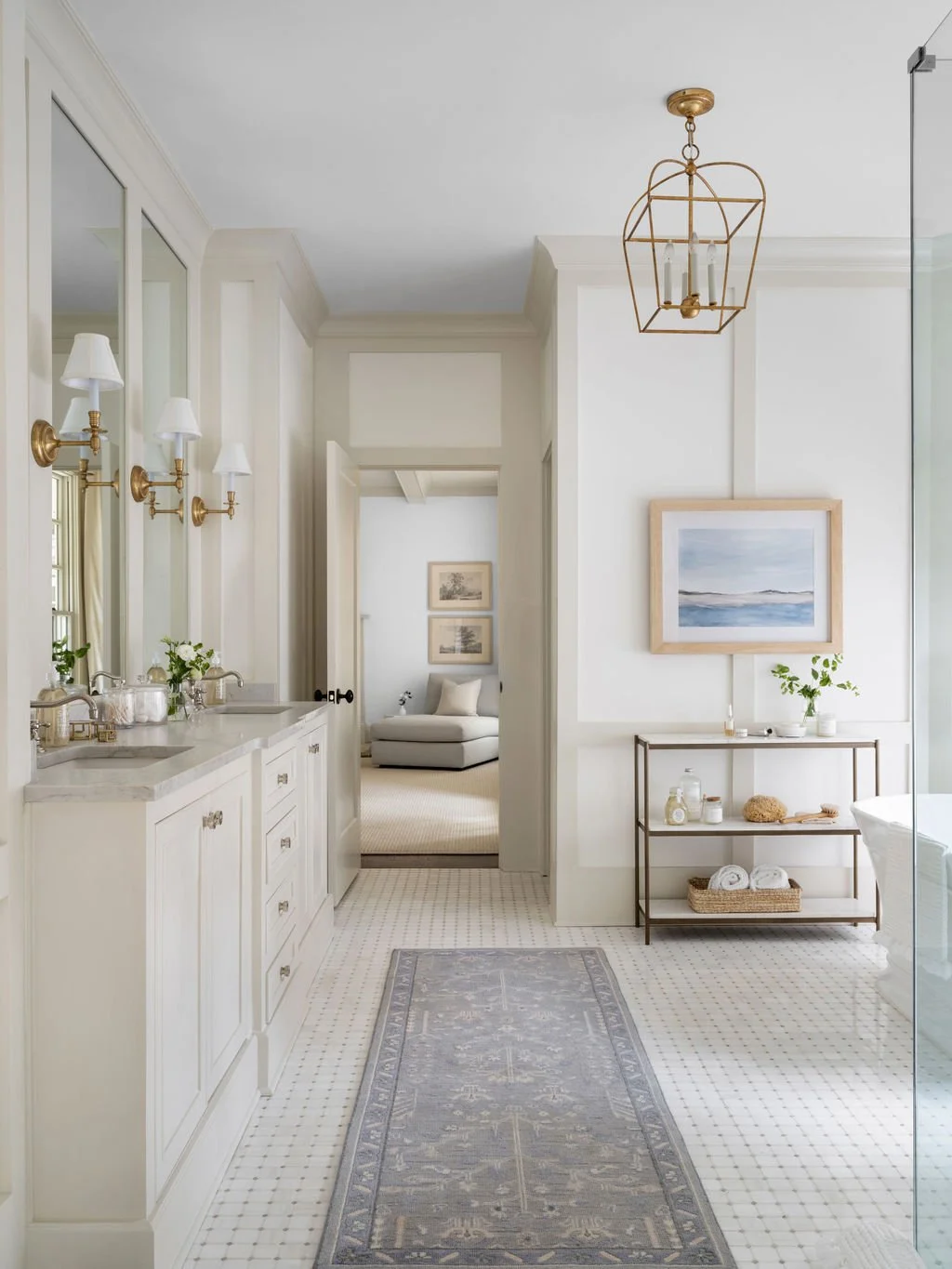 Bright bathroom with a double sink vanity, gold fixtures, and wall-mounted lamps. A rug on white tile flooring leads to a view into the adjacent sitting area with a gray chaise lounge and framed artworks. There is a metal shelving unit with towels, p
