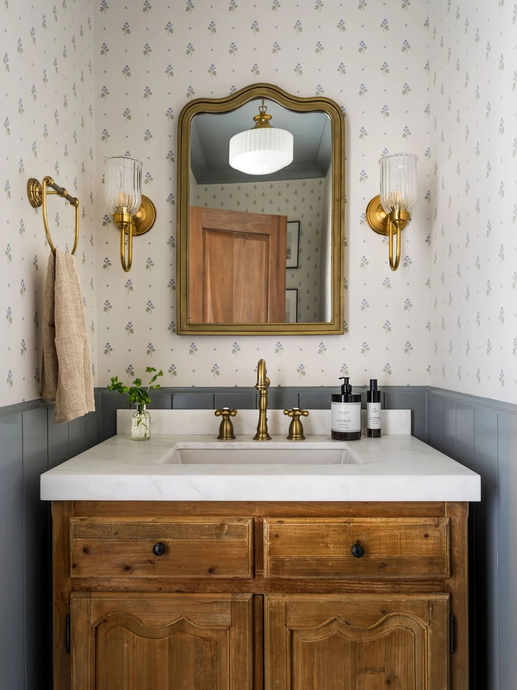 Bathroom vanity with a wooden cabinet, a white marble countertop, and a brass faucet. There is a mirror above the sink, flanked by two brass wall sconces with glass shades. A small plant and toiletries are on the counter, and a towel hangs on a brass