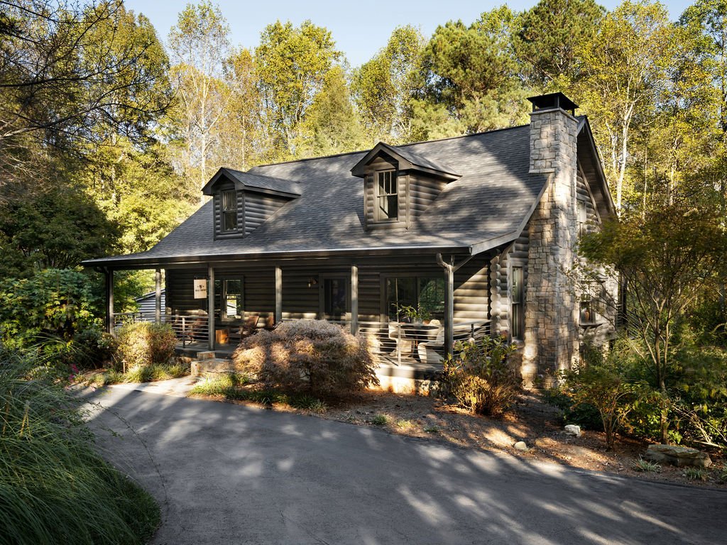 A log cabin-style house surrounded by trees and vegetation, with a curved driveway leading up to it, featuring a large stone chimney and a covered front porch with seating.