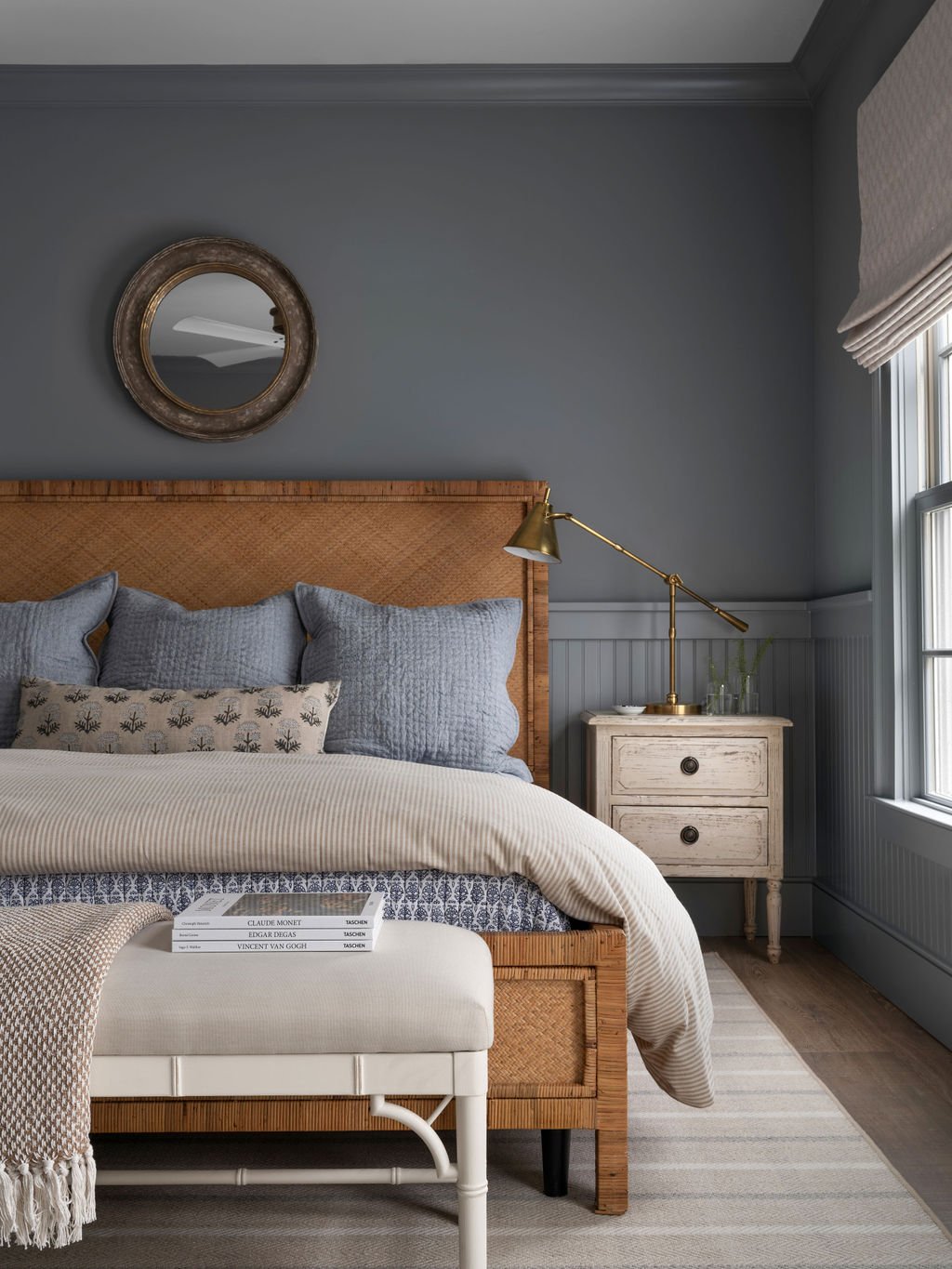 A cozy bedroom featuring a wooden bed with blue and patterned pillows, a white bench with books, a vintage white nightstand with a brass desk lamp, and a window with light-colored curtains, against gray walls and a striped rug.
