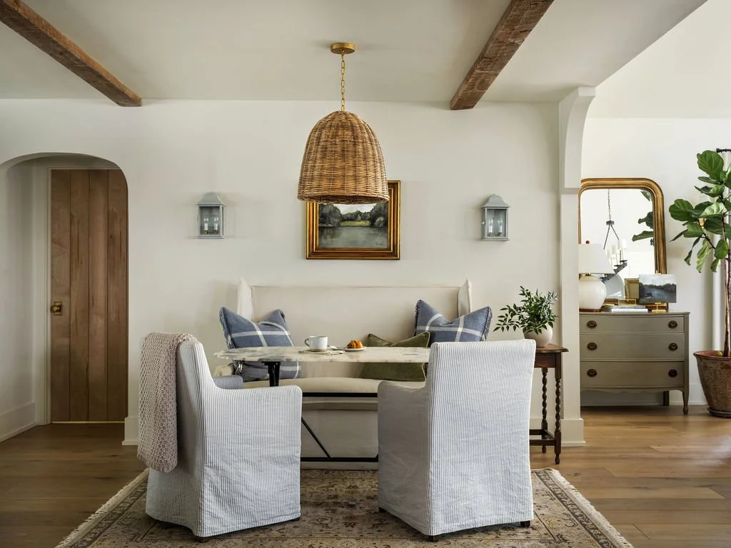 Cozy dining area with a white upholstered bench, two white armchairs, and a round table in a bright room with wooden beams on the ceiling, a framed landscape picture, a hanging wicker light, and a sideboard with decor and plants.
