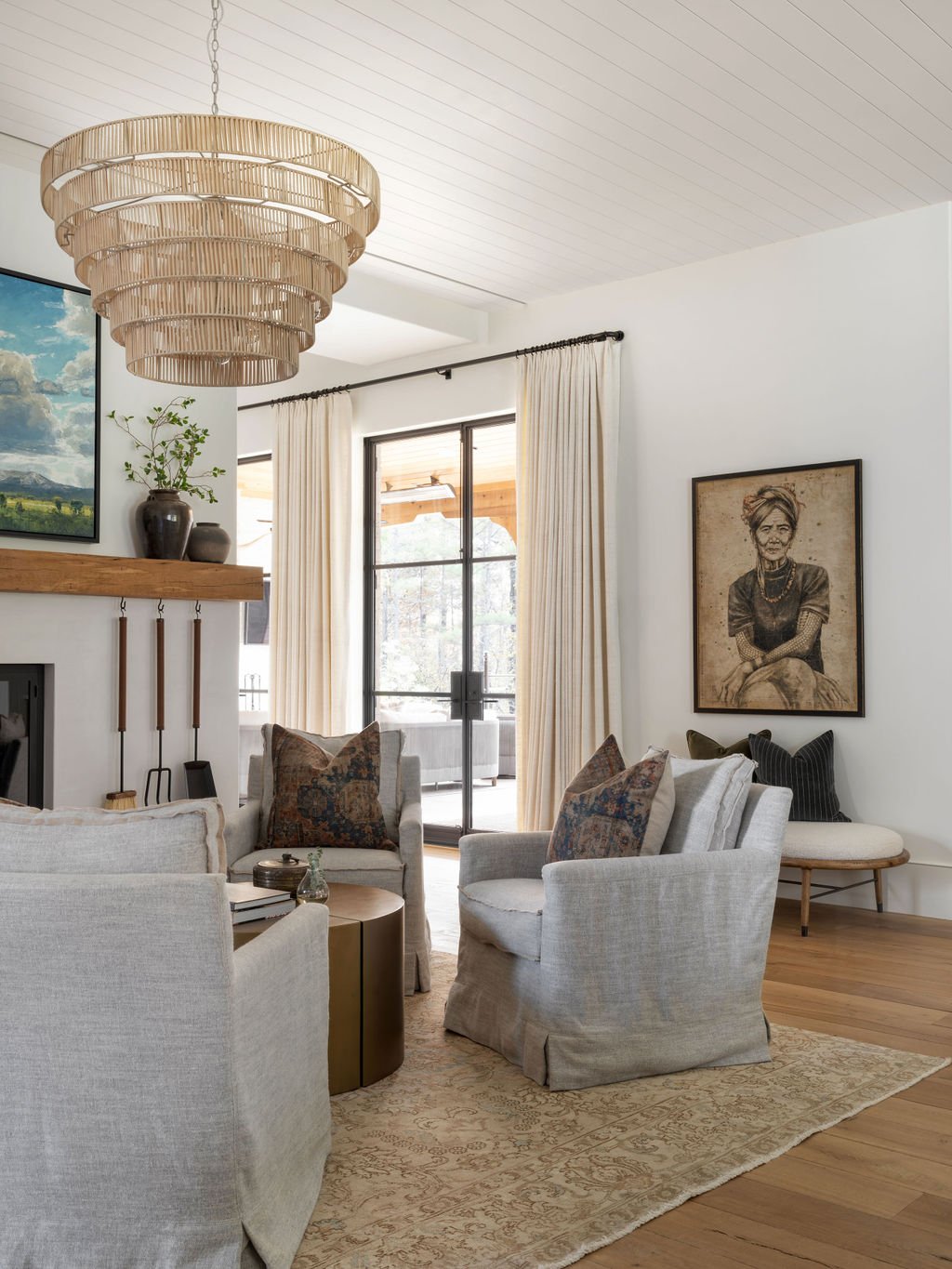 Living room with beige armchairs, a round coffee table, a decorative rug, and framed art on the wall. Large windows with cream curtains, a wooden mantel with vases and a plant, and a modern wooden chandelier.