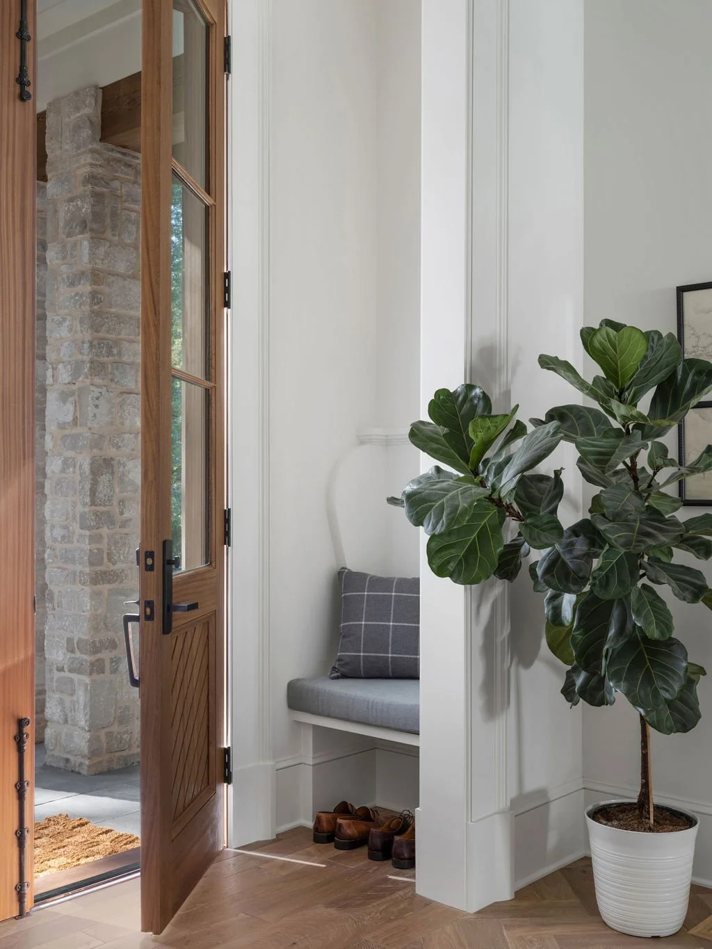 Entrance area with wooden door open to outside, stone wall visible through door, small bench with gray cushion and checkered pillow, three pair of shoes on the floor, large potted green plant, framed artwork on white wall.