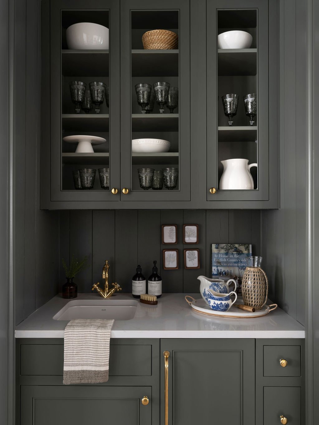 Dark gray kitchen cabinet with glass doors, display of white and black dishes, glasses, and a pitcher. White countertop with a sink, soap dispensers, decorative pitcher, and a tray with a woven vase and glassware.
