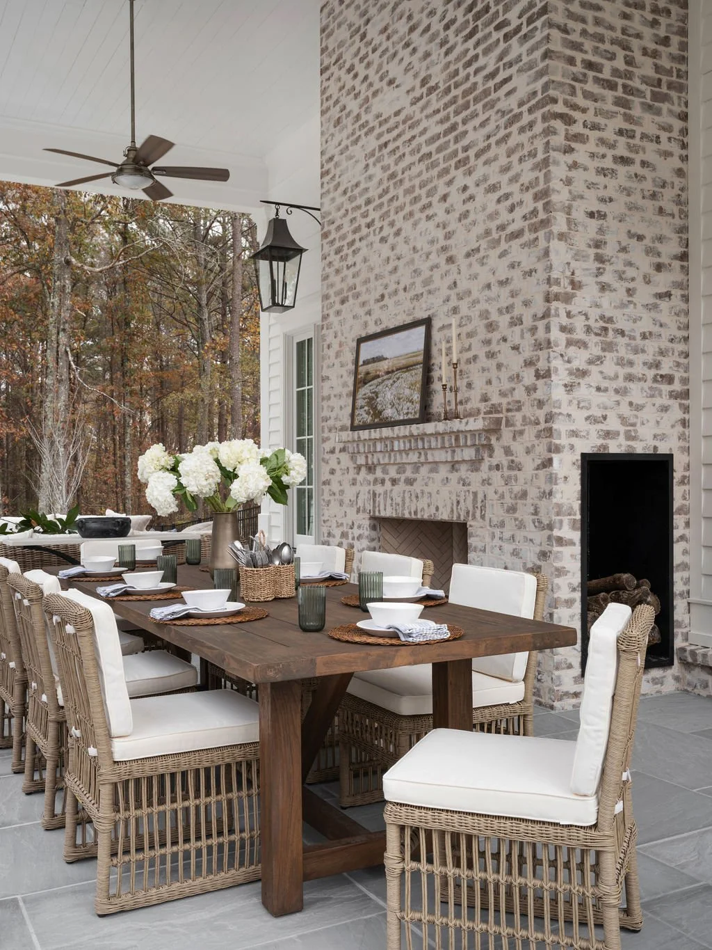 Outdoor dining area with a wooden table, white cushioned wicker chairs, a large vase with white hydrangeas, and a brick fireplace, under a ceiling fan and lantern, with trees in the background.
