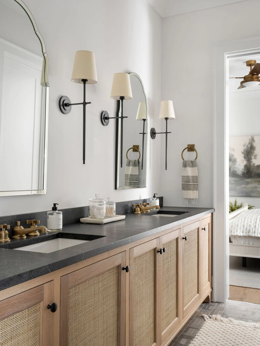 Bathroom with dual sinks, wooden cabinet, wall-mounted lamps, mirror, towel ring, and toiletries, near a bedroom with a ceiling fan and landscape painting.