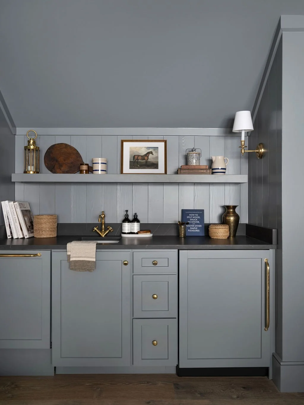 A kitchen with light blue cabinets, black countertop, and gray wood-paneled wall. Decor includes a framed horse picture, a golden vase, books, baskets, and kitchen accessories.