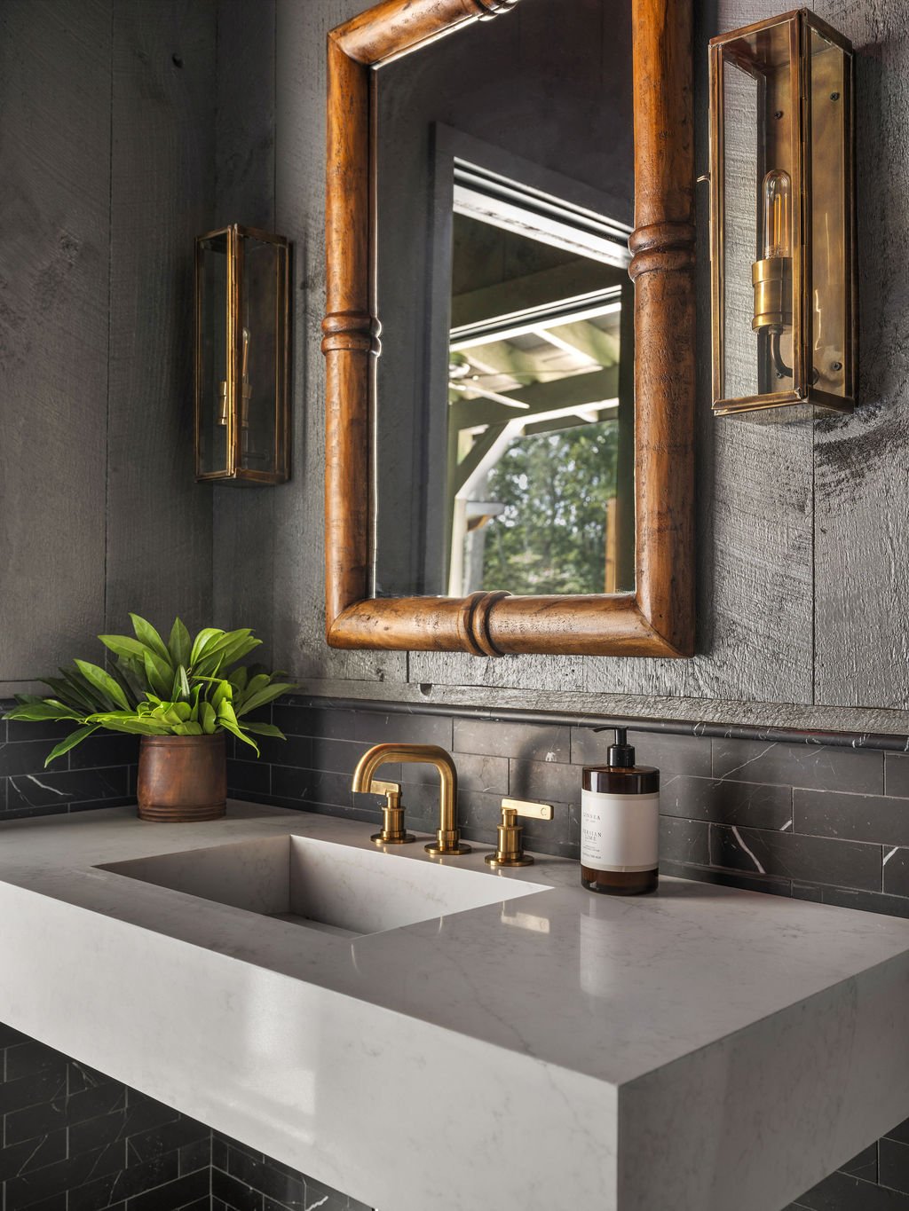 Modern bathroom with a dark wall, large wooden-framed mirror, gold fixtures, a white sink, potted green plant, and wall-mounted brass light fixtures.