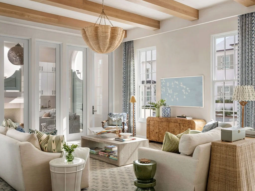 Bright living room with white walls, large windows, and wooden ceiling beams. Features a white sofa with patterned cushions, a marble coffee table, and various decorative items. An open door leads to an outdoor patio area with seating.