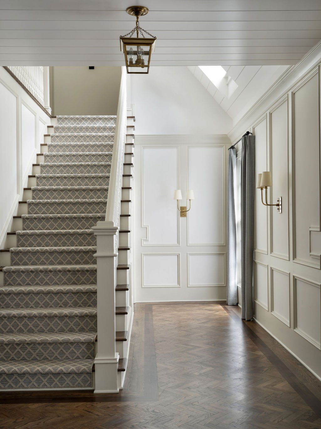 Interior view of a foyer with a staircase carpeted in a diamond-patterned runner, wooden steps, white wainscoting, a hanging lantern, wall sconces, and a window with curtains.