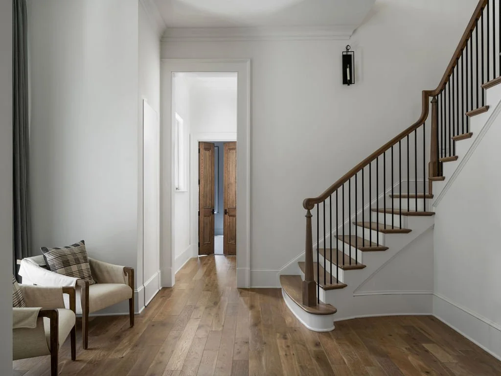 Empty foyer with wooden staircase on the right, a small bench with plaid pillows on the left, light hardwood floors, white walls, a door at the end of the hallway, and a hanging light fixture.