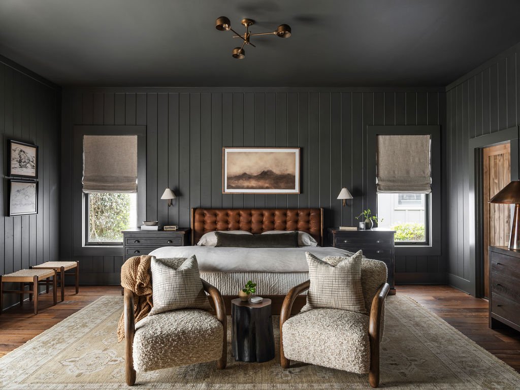 A bedroom with dark gray walls, a brown tufted headboard, two nightstands with white lamps, two small windows with gray blinds, two armchairs with pillows, a rug, and a ceiling light fixture.