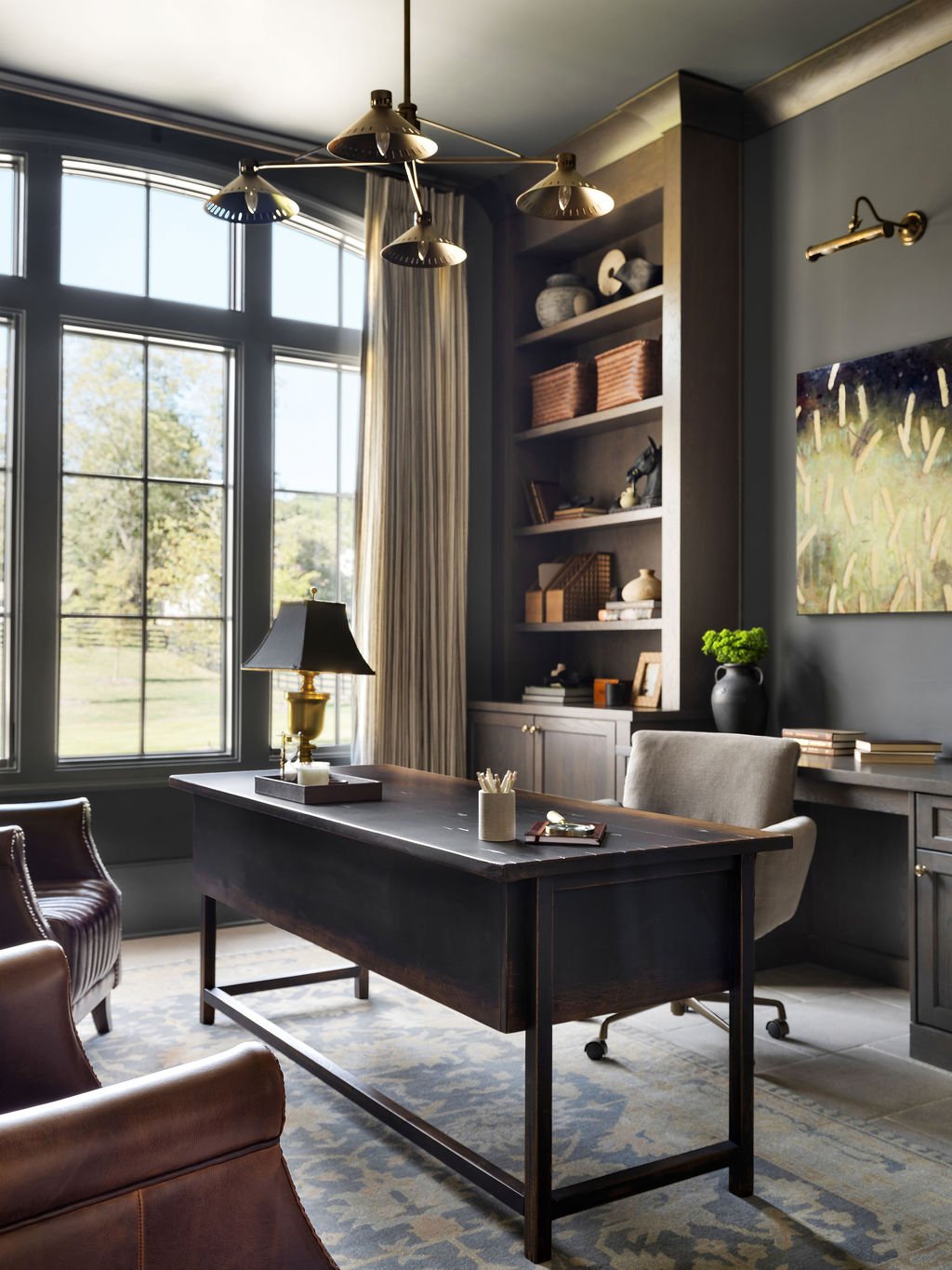 A cozy home office with a large window, dark wooden desk, beige office chair, and shelves filled with decor and books. Natural light illuminates the room.