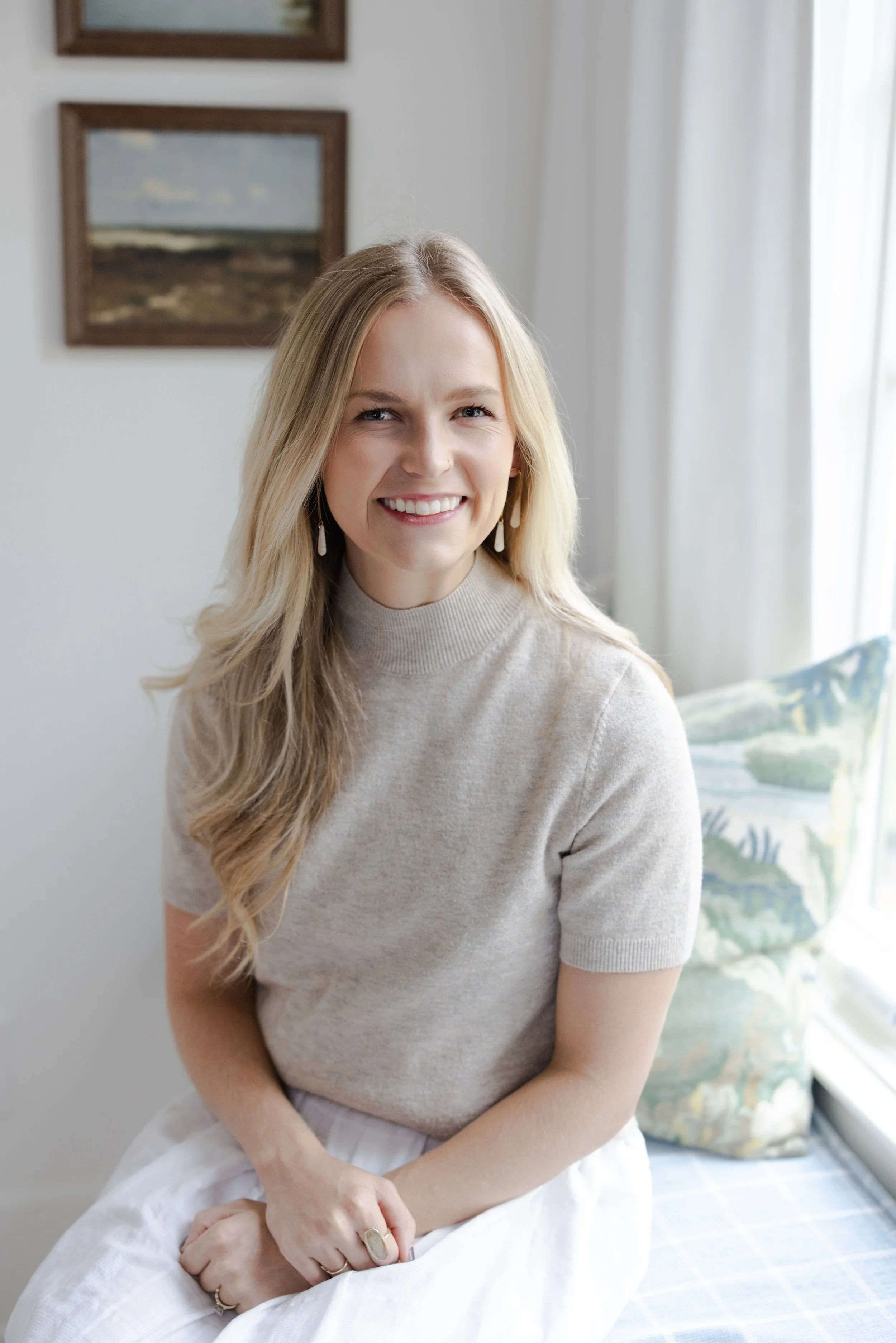 A smiling young woman with long blonde hair, wearing a beige short-sleeved sweater and white skirt, sitting on a window seat with a patterned cushion, inside a room with framed landscape paintings on the wall.
