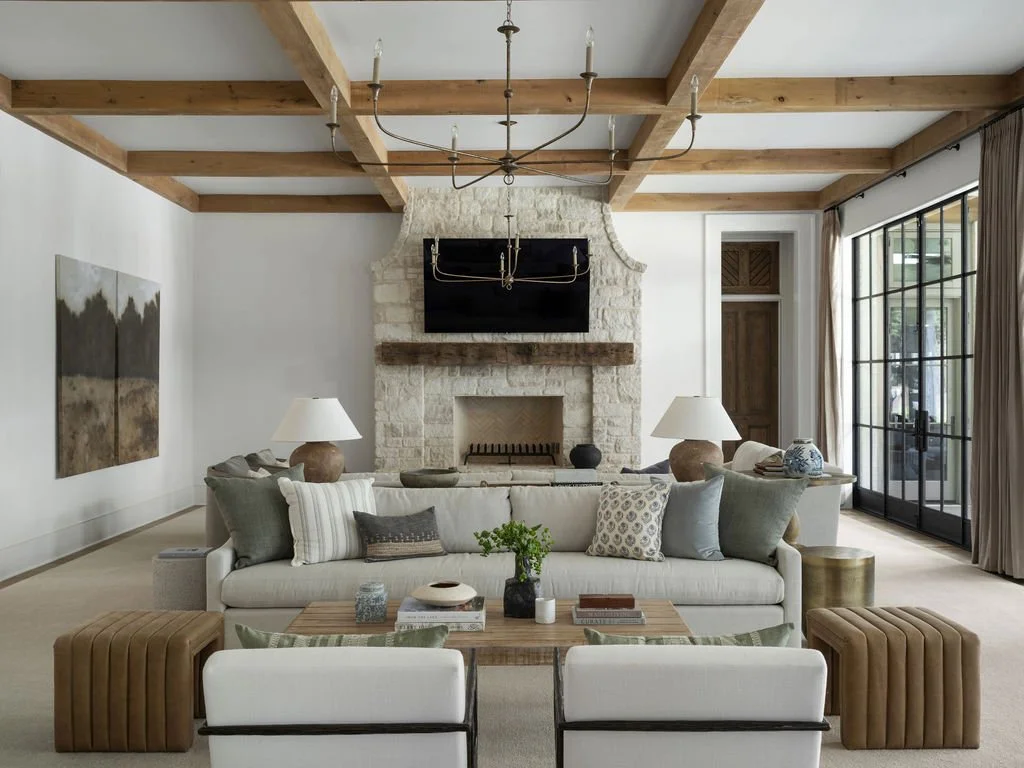A living room with a white sofa, decorative pillows, two lamps, a wooden coffee table with books and decor, leather ottomans, a stone fireplace with a mounted TV, and large windows with curtains.