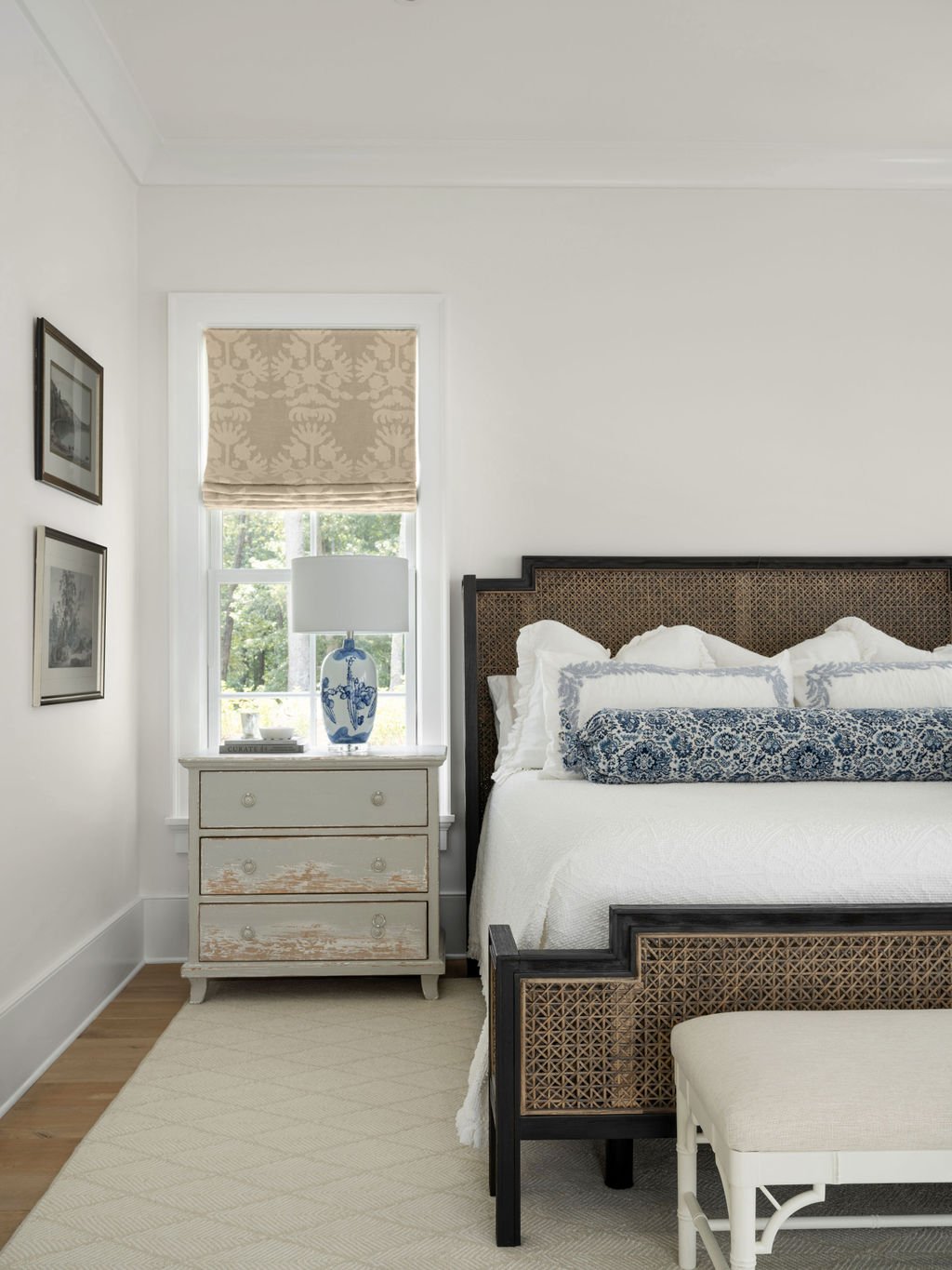 A bedroom with a white bed, a wicker headboard, and pillows, next to a white nightstand with a blue and white lamp, window with a beige patterned shade, and framed pictures on the wall.