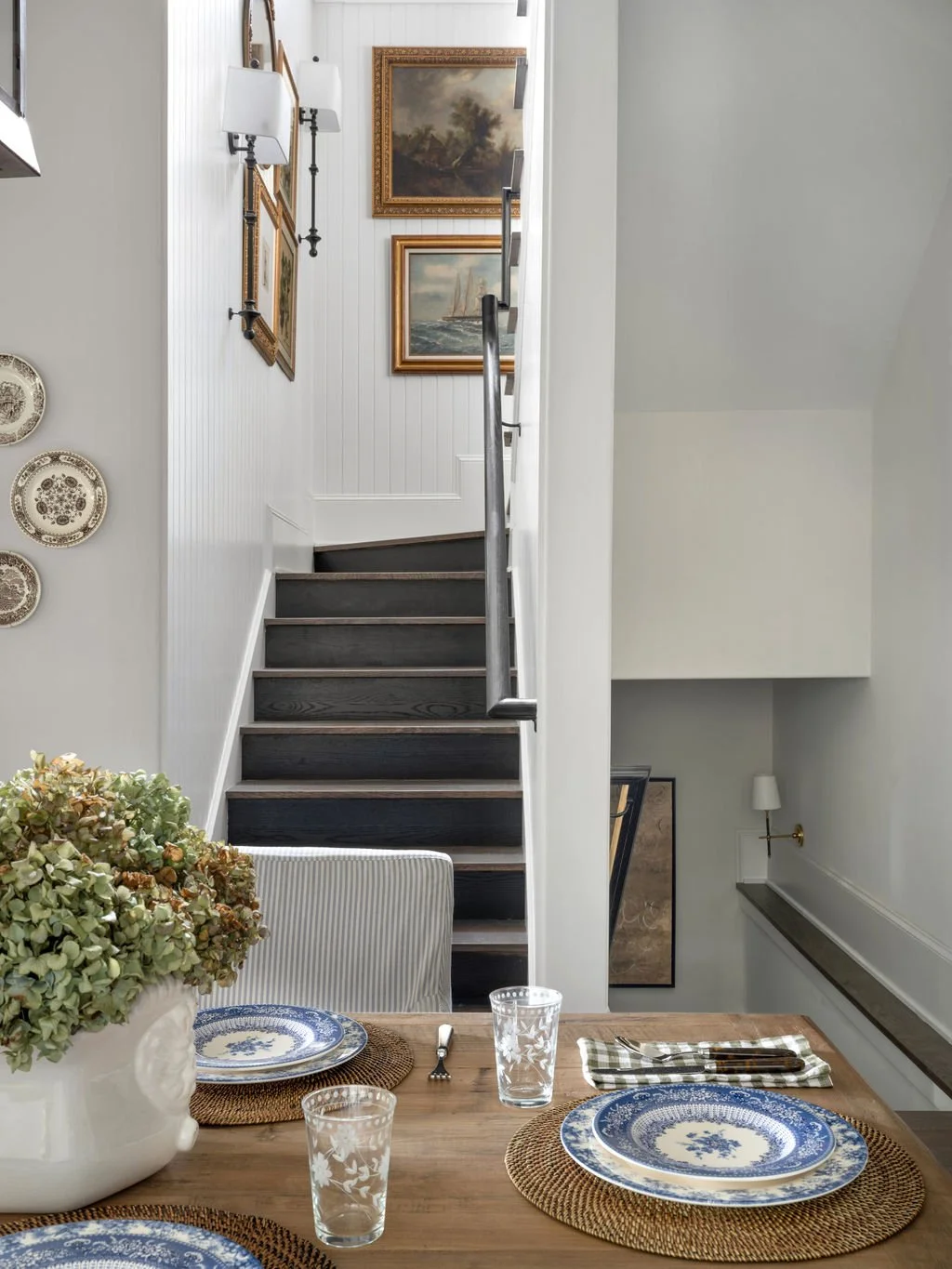Interior view of a dining area with a wooden table, blue and white patterned plates, glasses, and a floral centerpiece, with a staircase and framed paintings on the white paneled wall in the background.