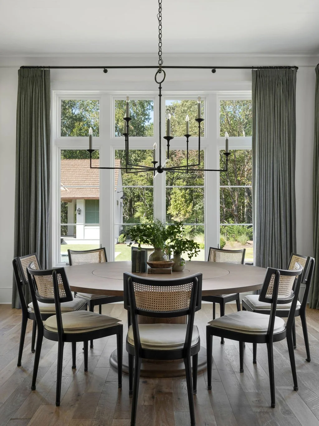Dining room with a large window, round table with chairs, chandelier, and a vase with greenery.