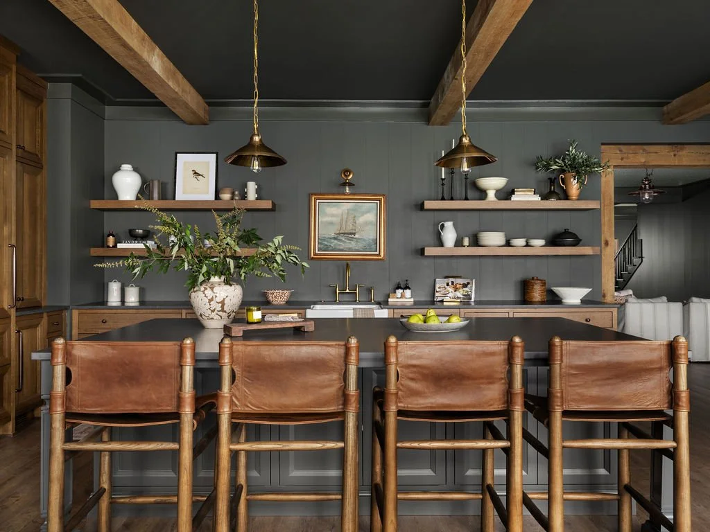 A modern kitchen with a dark gray wall, wooden beams on the ceiling, and open wooden shelves displaying dishes, bowls, and decorative items. There is a large central island with a dark countertop, surrounded by four wooden chairs with tan leather sea