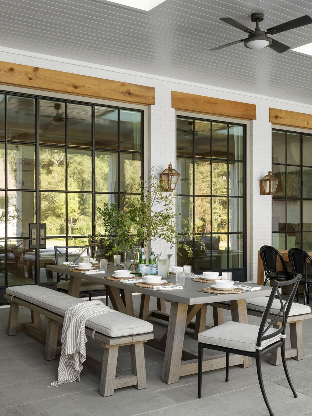 Outdoor dining area with a wooden dining table set with white bowls, glasses, utensils, and a centerpiece with green foliage, surrounded by chairs and a long bench, with a white brick wall and large glass doors behind.