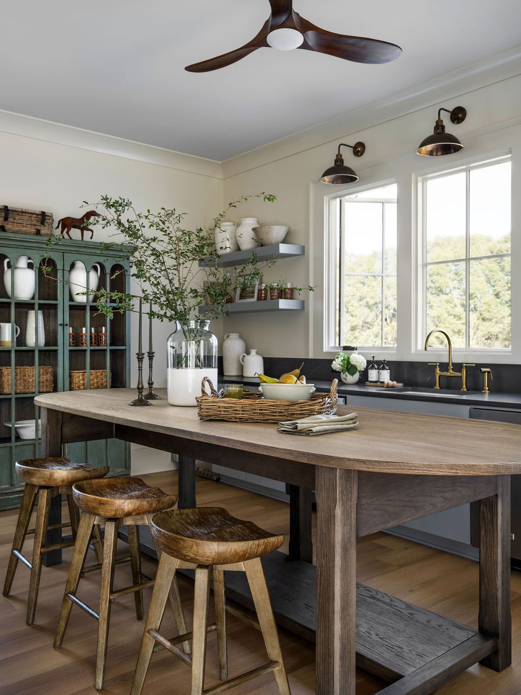 A rustic kitchen with a wooden dining table, three matching wooden stools, a green cabinet with white and brown ceramics, a large glass jar with greenery, a woven tray with fruit, and a window with a view of trees outside.